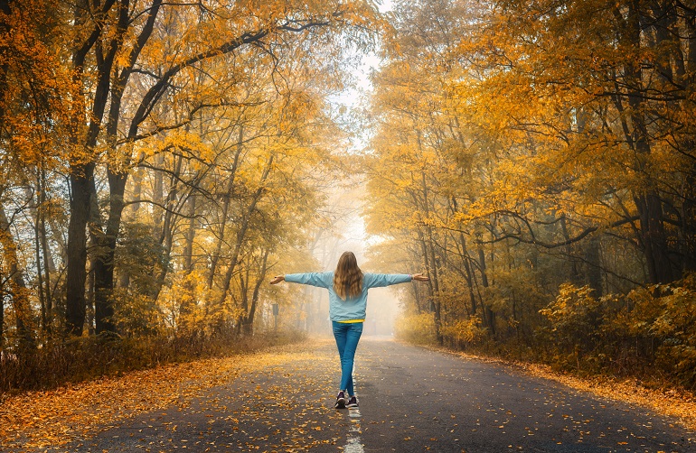Happy young woman on the road in beautiful autumn forest in fog at sunset. Landscape with alone girl, road, orange trees. Empty roadway through foggy woodland in fall. Travel and adventure in Europe